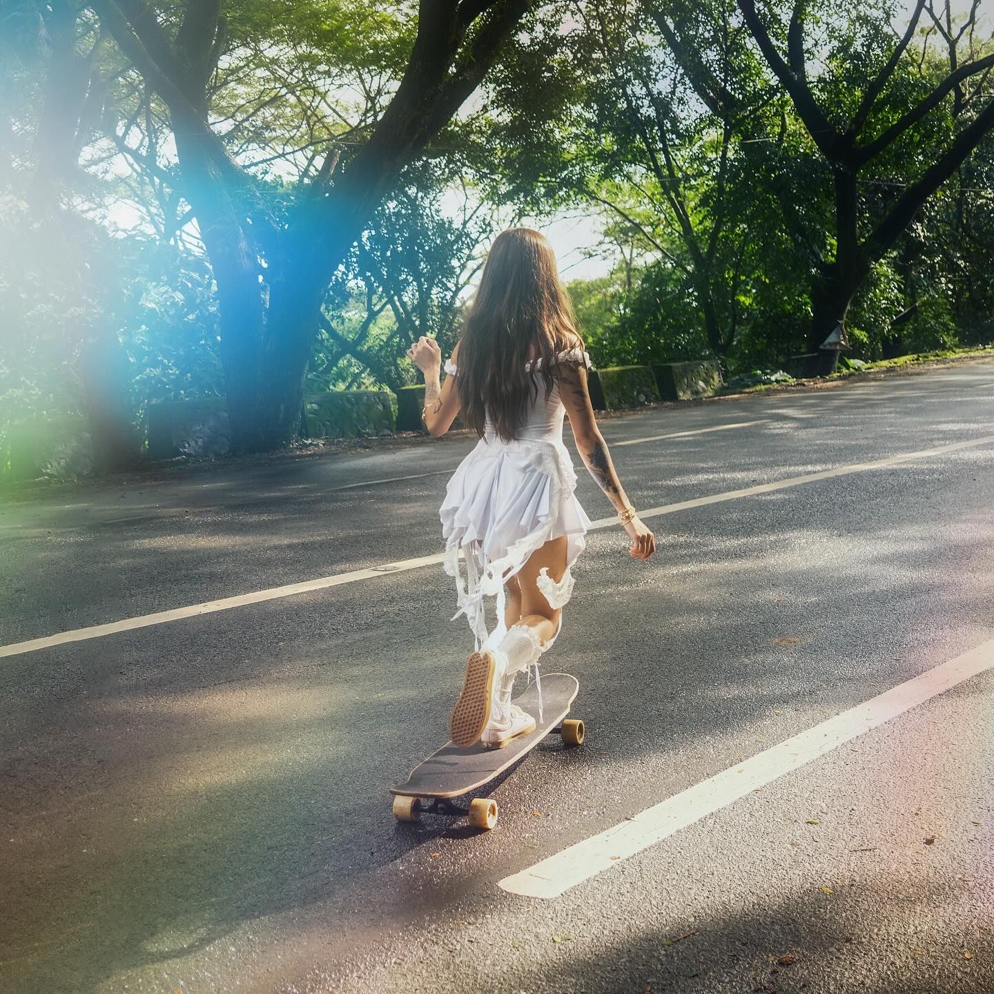 The image depicts syd hartha while skateboarding by the road, with trees in the surrounding.